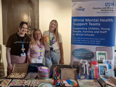 A picture of 3 staff members wearing lanyards standing in front of a table full of leaflets and craft materials.The sign beside them says 