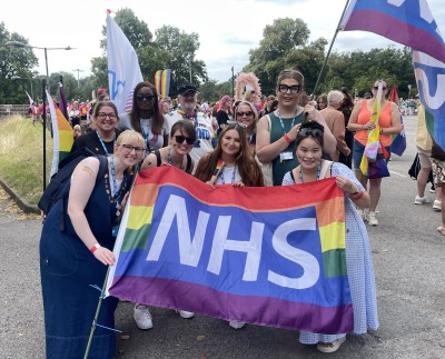 A picture of 9 staff members wearing lanyards. They are outside, holding large rainbow flags that have the NHS logo on them. 
