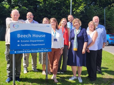 A picture of 8 people. Isla Wilson, CWP Chair, at Beech House in Macclesfield. Meeting with our CYP Autism and ADHD Service team. They are behind an NHS sign that says 
