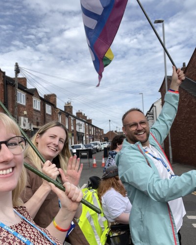 A picture of 3 staff members wearing lanyards. They are holding flags in a parade.