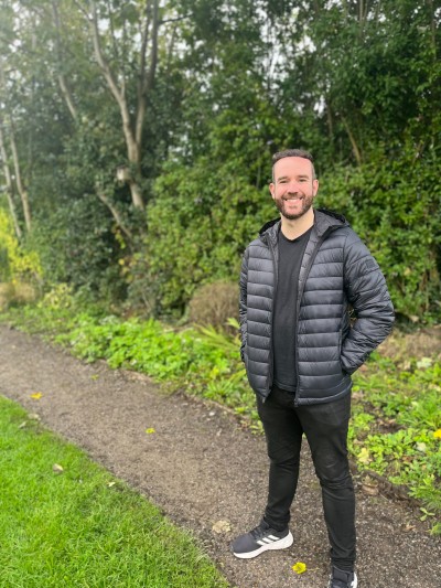 A person named Rich from this patient story is stood in front of green trees smiling at the camera